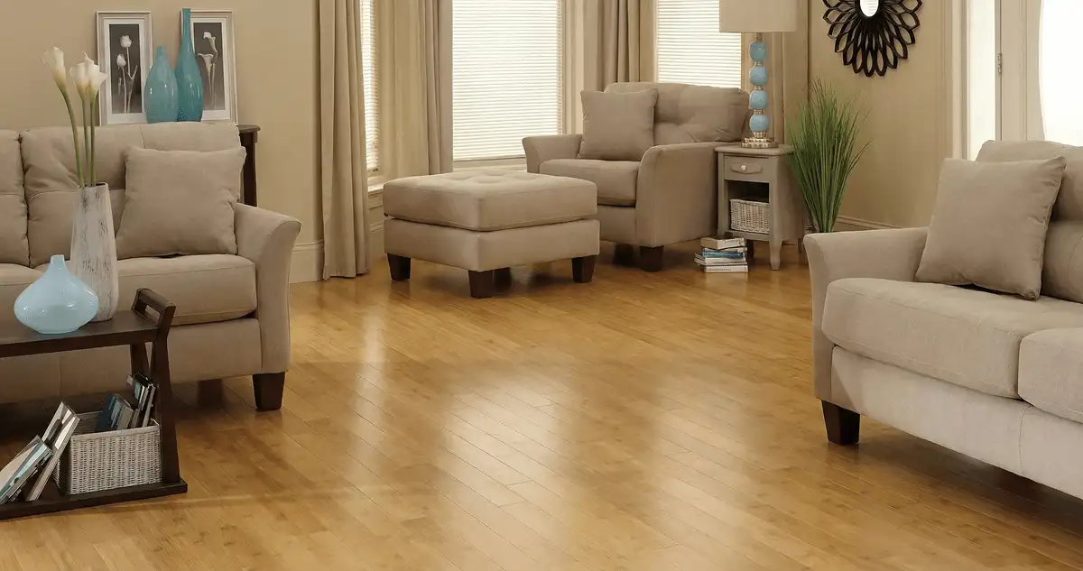 A living room with professionally installed bamboo flooring, beige sofas, a matching ottoman, a side table with a blue lamp, vases, framed pictures, and large windows with blinds and curtains in Pembroke Pines, FL